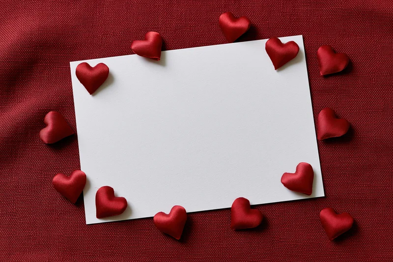 Blank white card surrounded by red satin hearts on red fabric surface