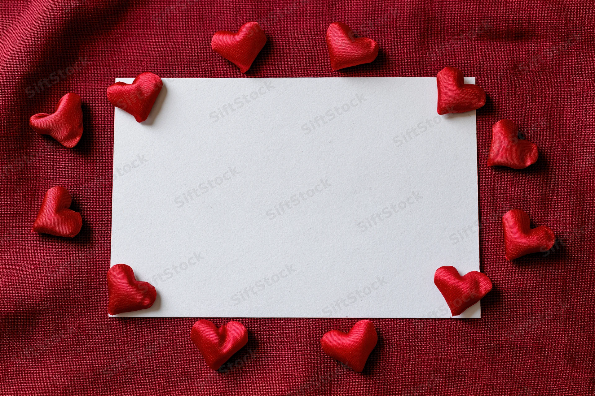 Blank white card surrounded by red hearts on red fabric surface