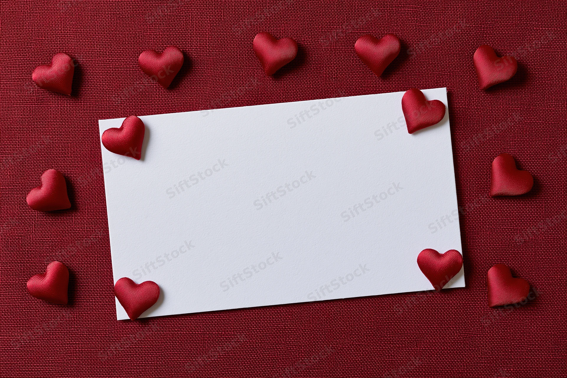 Blank white card surrounded by red hearts on textured red fabric surface