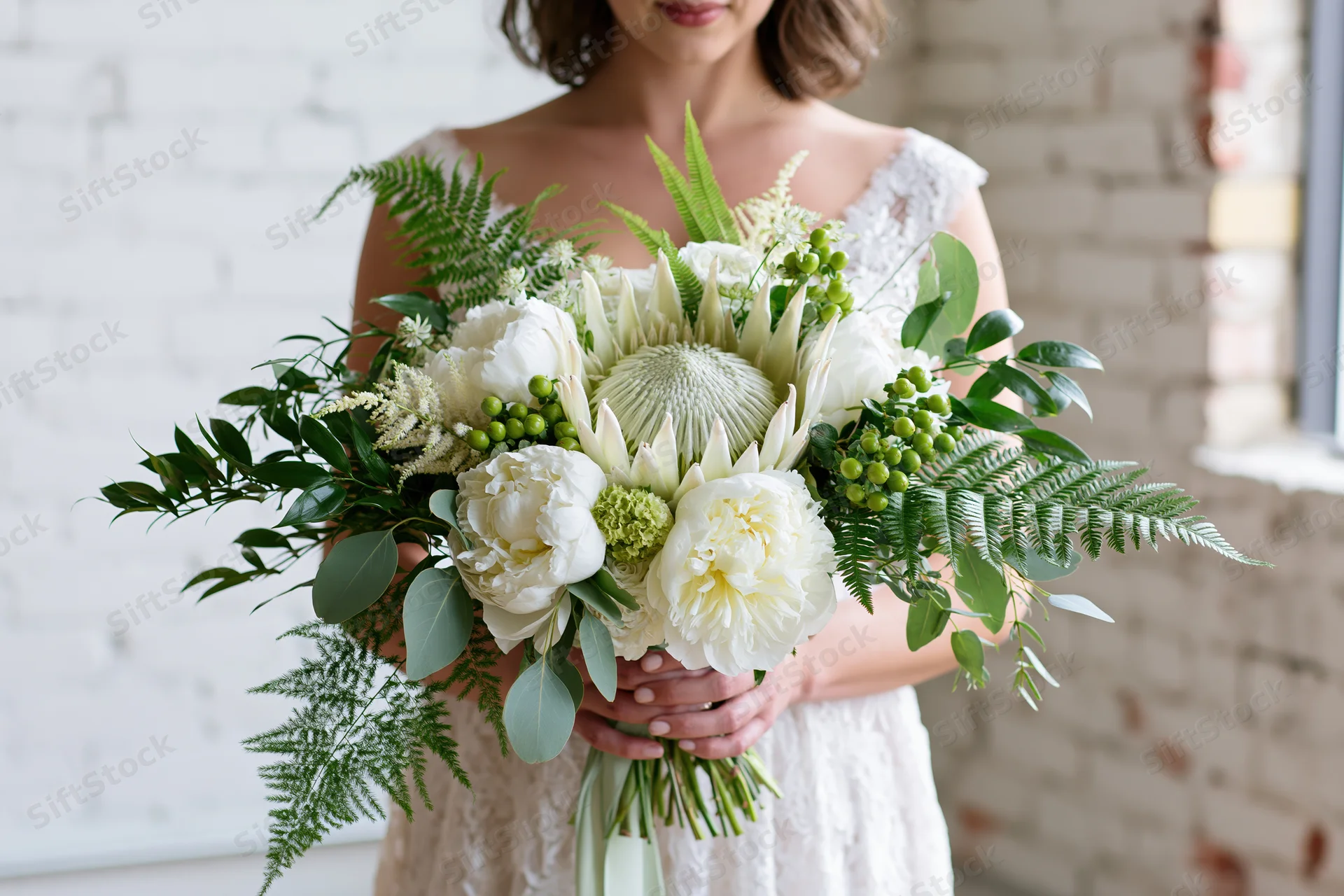 Bride holding a large cascading bridal bouquet with white peonies and green foliage indoors