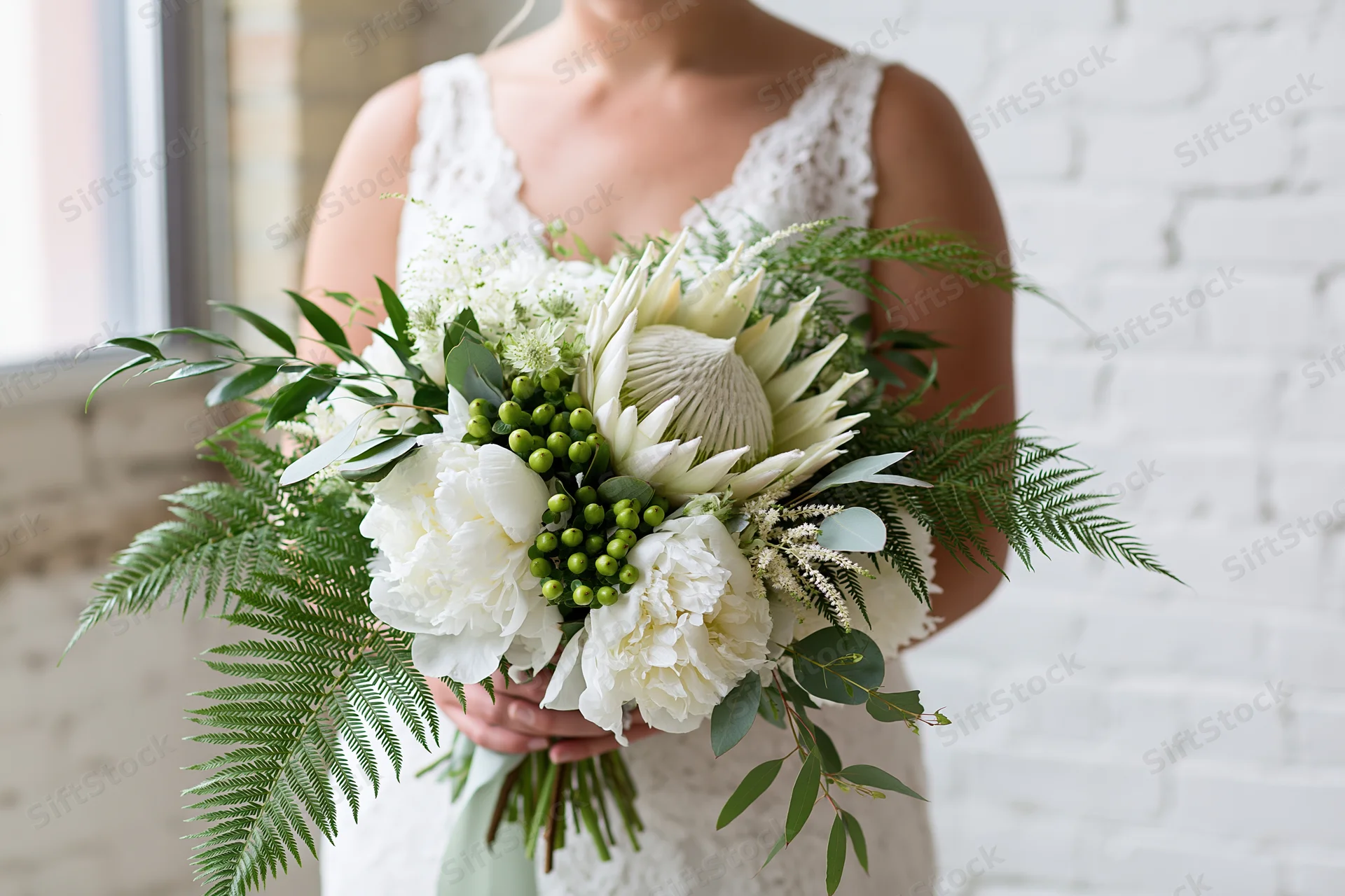 Bride holding a cascading bridal bouquet of white peonies and green ferns against a white brick wall