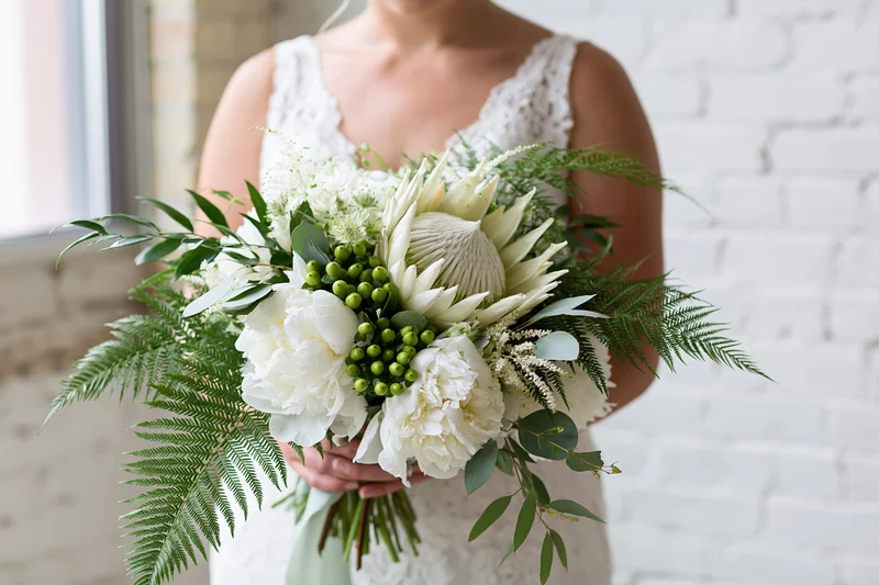Bride holding a cascading bridal bouquet of white peonies and green ferns against a white brick wall
