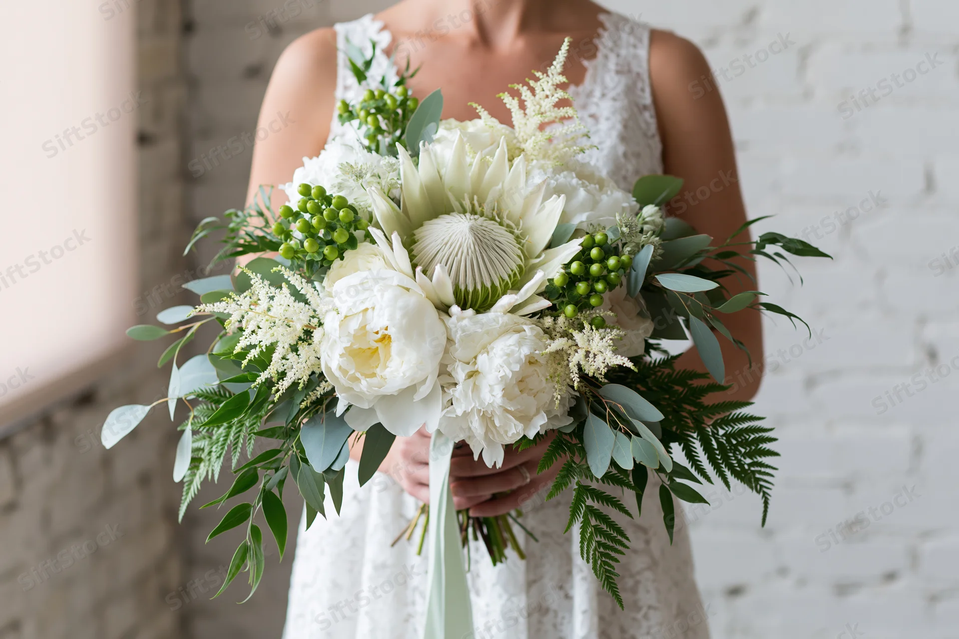 Bride holding a large cascading bridal bouquet with white peonies and green foliage against a white brick wall
