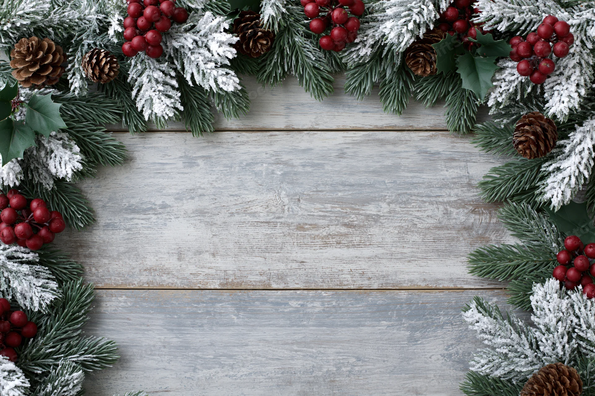Snow-dusted fir branches with pinecones and holly berries arranged as a border on a rustic wooden tabletop