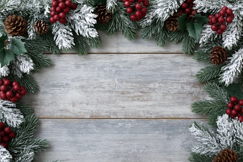 Snow-dusted fir branches with pinecones and holly berries arranged as a border on a rustic wooden tabletop