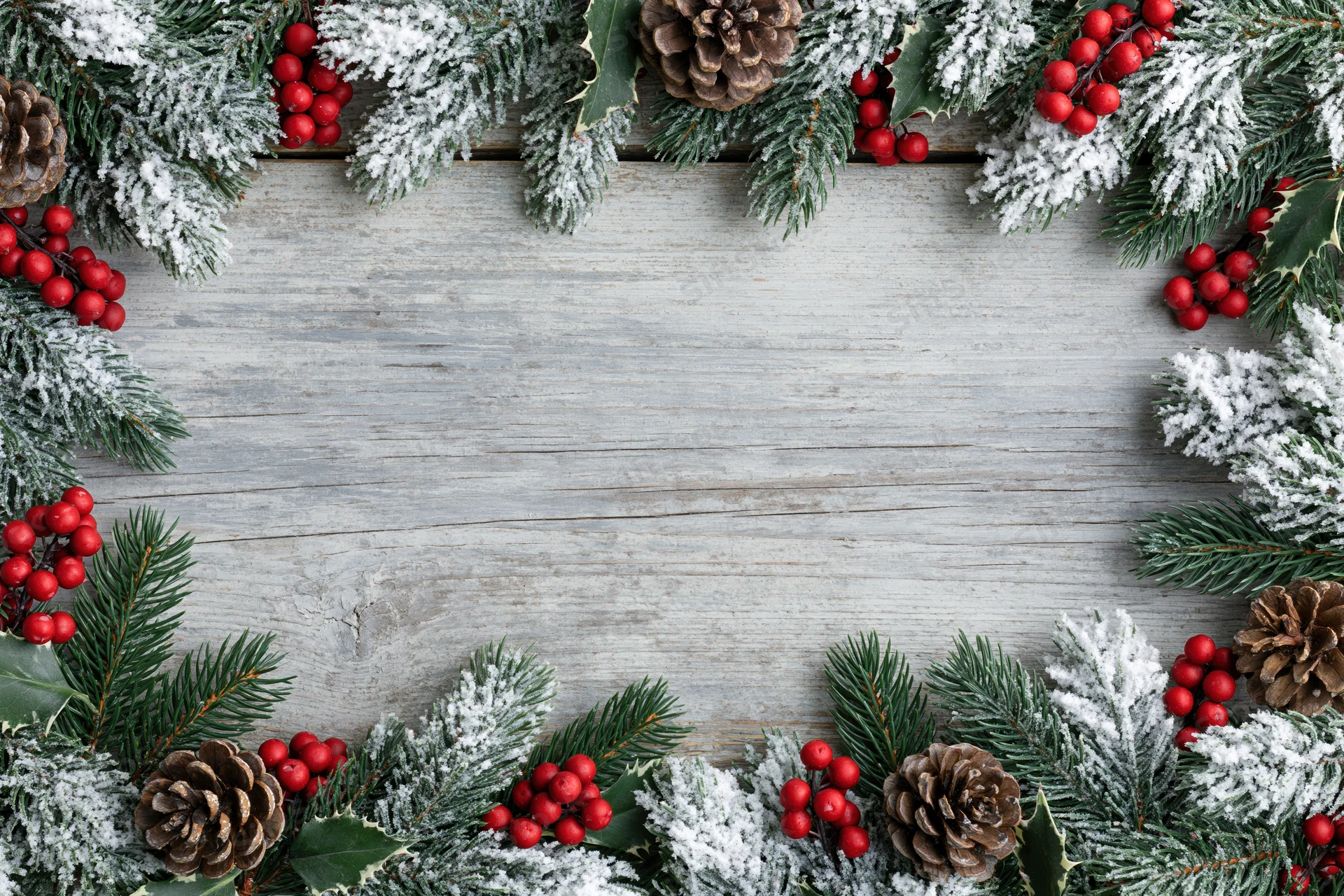 Snow-dusted fir branches with pinecones and holly berries arranged in a border on a rustic wooden surface