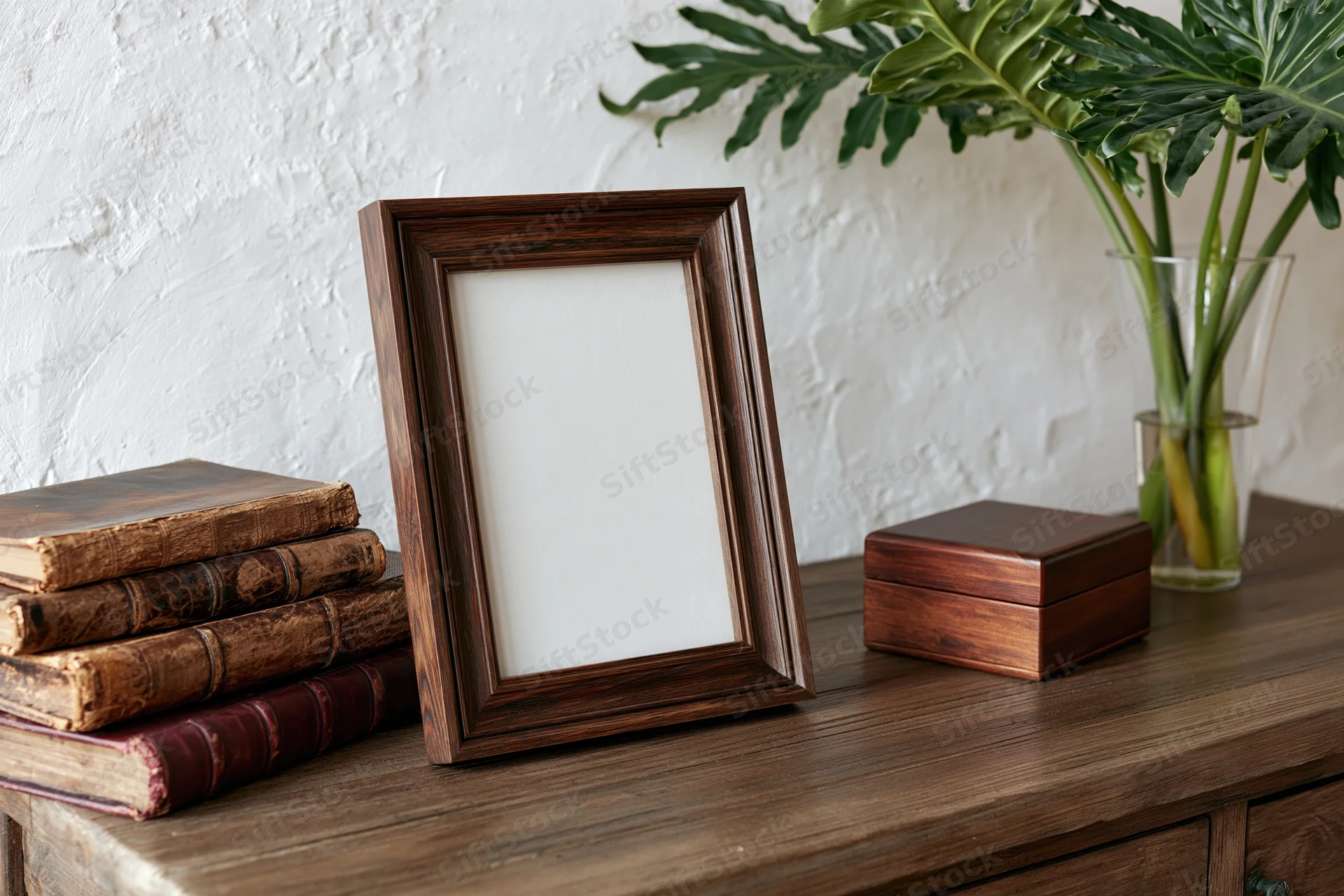Dark wood picture frame with blank white insert leaning on wooden cabinet beside stack of vintage books and potted monstera plant