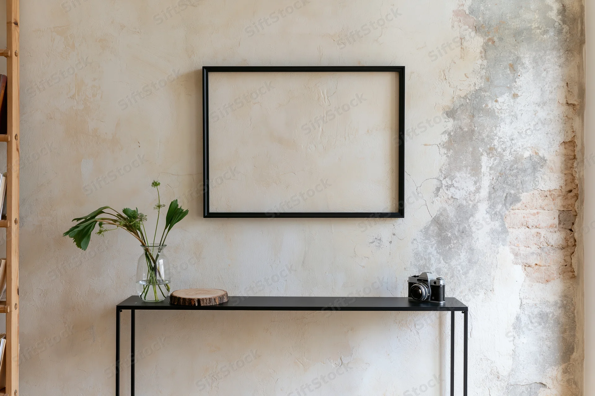 Empty black frame hanging on weathered plaster wall above metal console table with vase of green stems and vintage camera