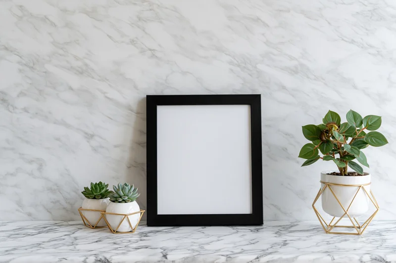 Empty black picture frame leaning against white marble wall with potted plants on countertop