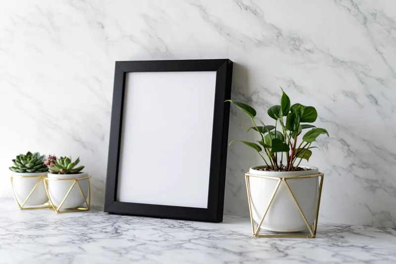 Empty black picture frame leaning against marble wall beside potted plants