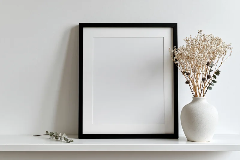 Empty black frame with white mat leaning on white shelf beside ceramic vase holding dried flowers
