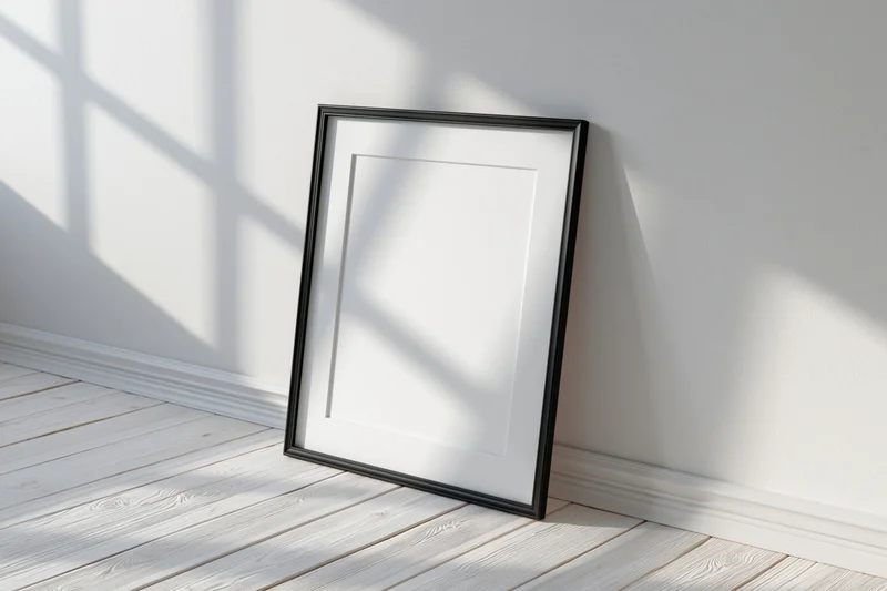 Black picture frame with white mat leaning against white wall on wooden floor in bright sunlight