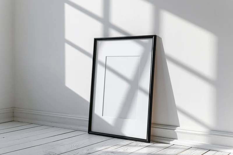 Black picture frame with white mat leaning against white wall in sunlit corner of minimalist room