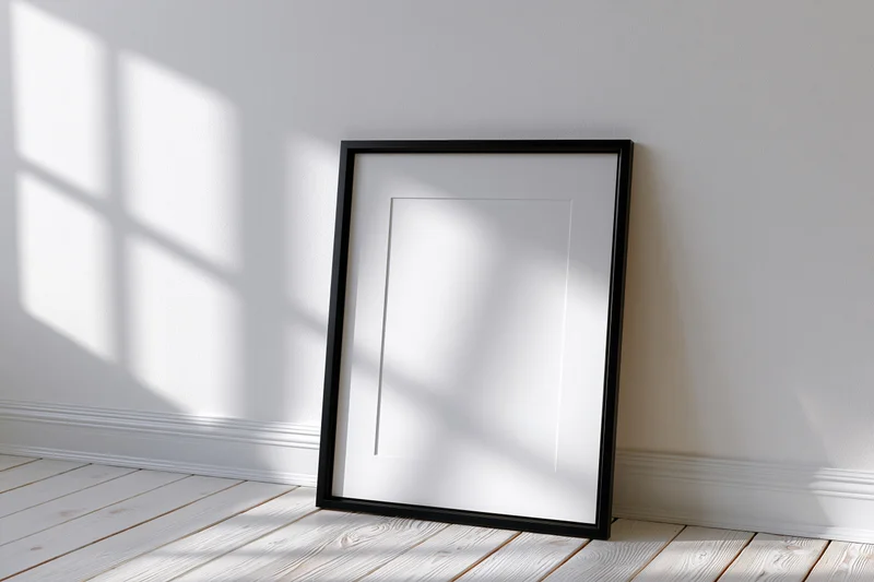 Black picture frame with white mat leaning against white wall on wooden floor in sunlit room