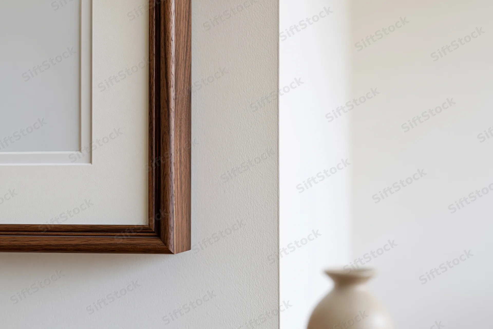 Dark walnut wood picture frame with white mat board hanging on a white wall beside a blurred ceramic vase