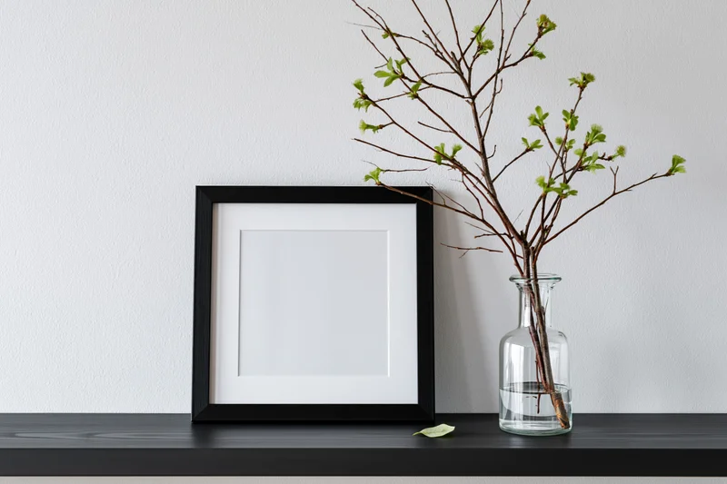Empty black picture frame with white mat leaning on dark shelf beside glass vase holding spring branches