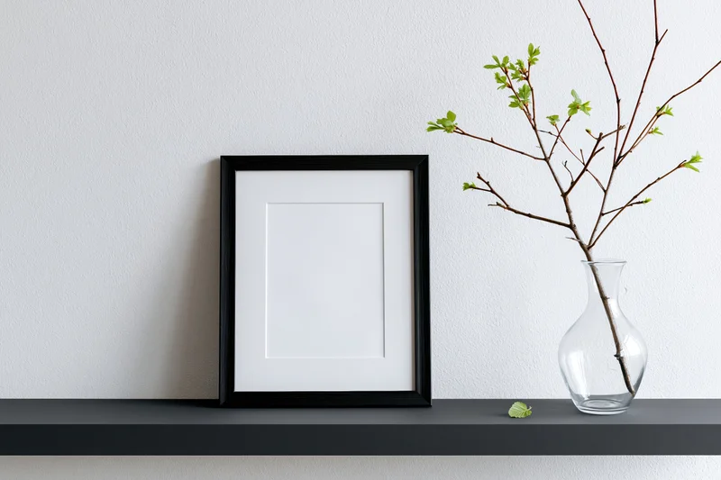 Black picture frame with white mat leaning on dark shelf beside glass vase holding spring branches