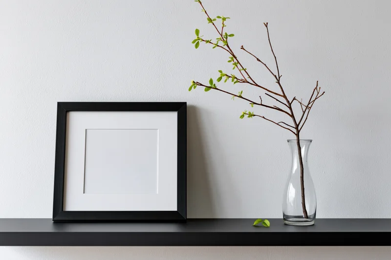 Empty black picture frame with white mat leaning on black shelf beside glass vase holding leafy branch