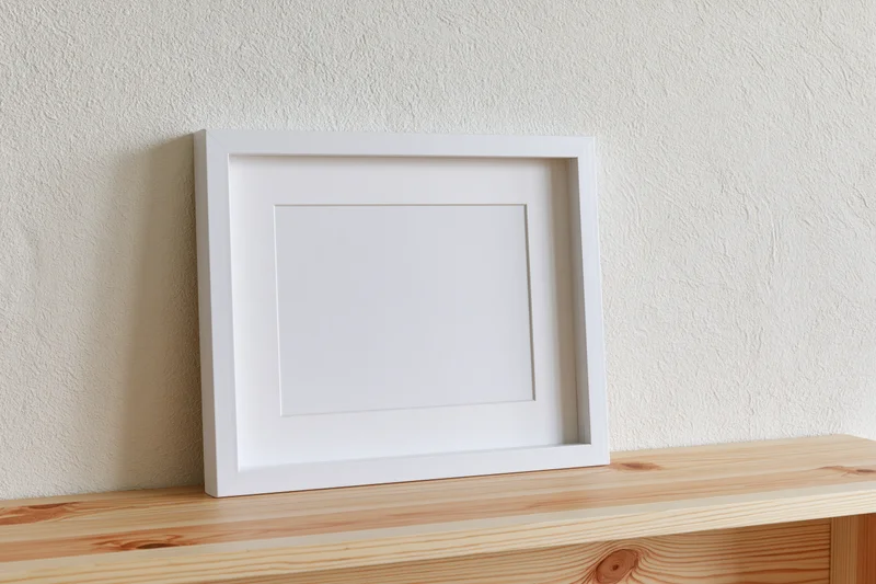 White empty picture frame leaning on wooden shelf against textured wall