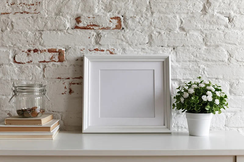 Empty white picture frame leaning on a shelf against a whitewashed brick wall with books, coins in a jar, and a potted plant