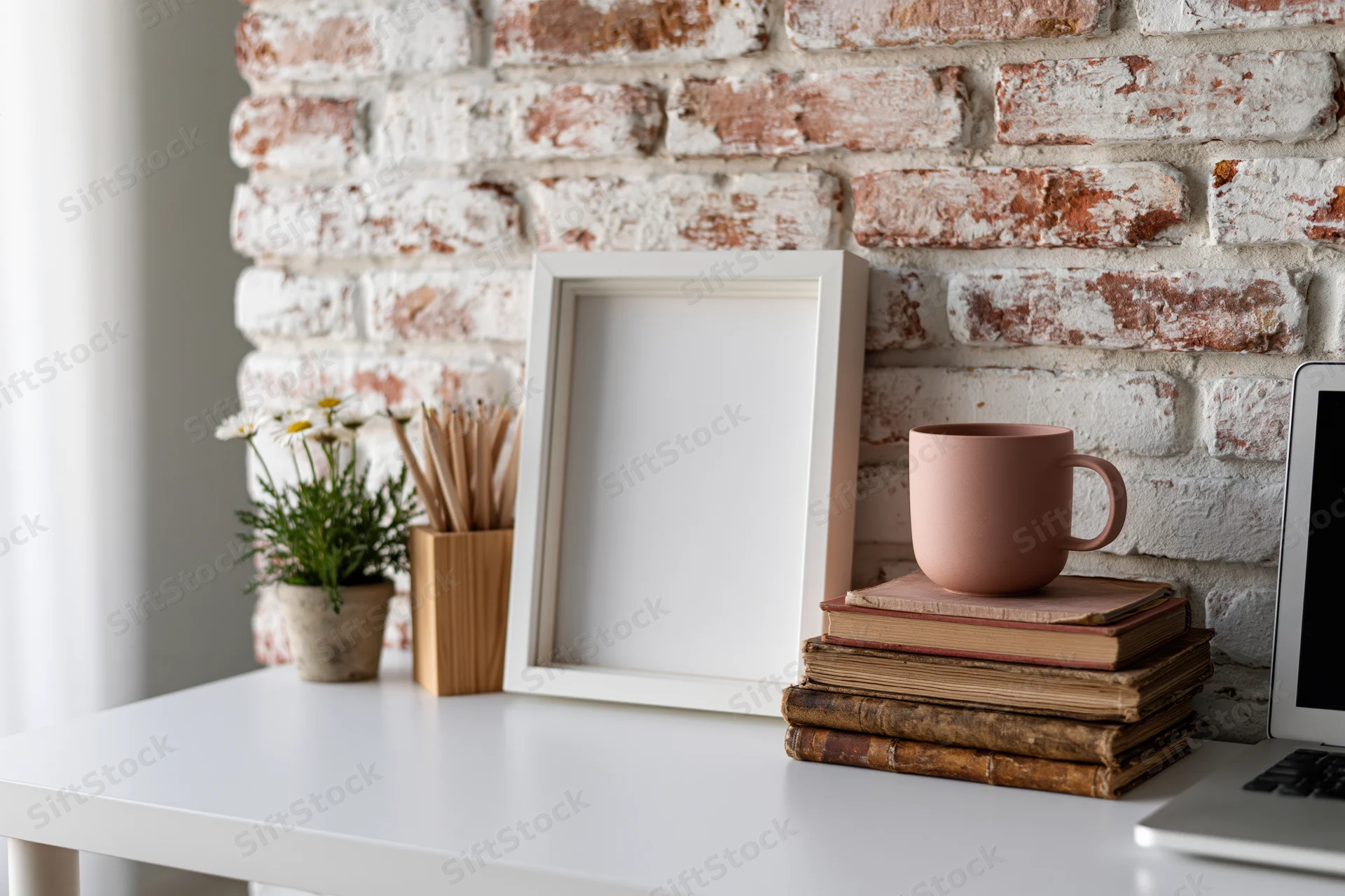 Empty white frame leaning on desk beside books and mug against whitewashed brick wall