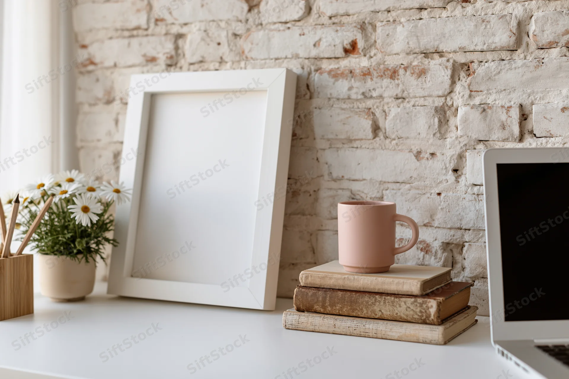 Empty white frame leaning against whitewashed brick wall on desk with laptop, books, mug and daisies