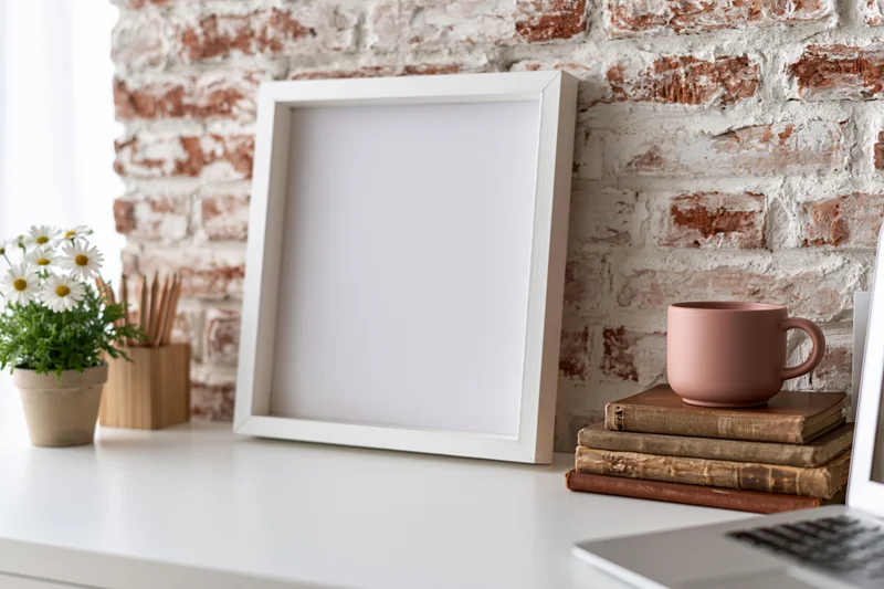 Empty white frame leaning on desk beside books and mug against whitewashed brick wall