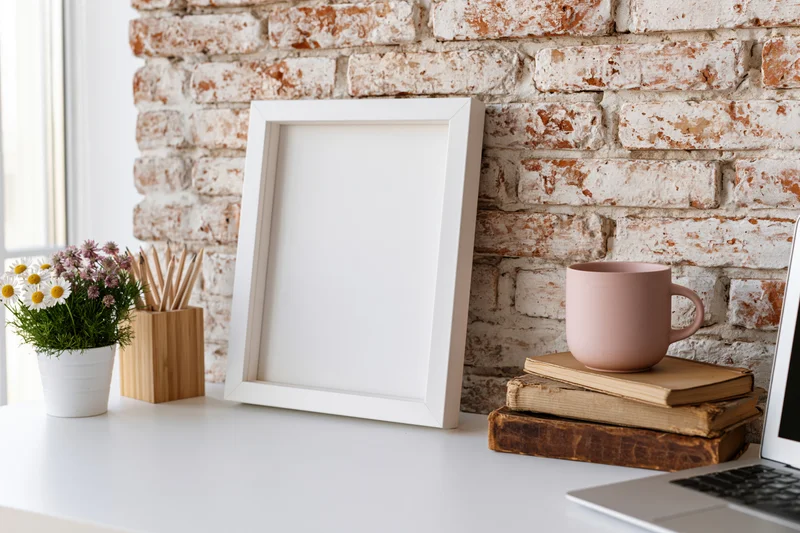 Empty white frame leaning on desk beside laptop and books with flowers and pencils in bamboo holder against whitewashed brick wall