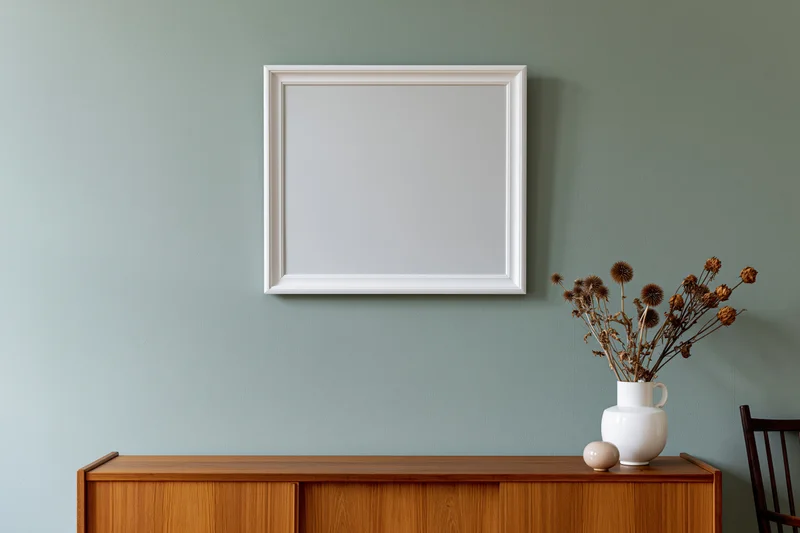 Empty white frame hanging on a sage-green wall above a wooden console with a vase of dried flowers