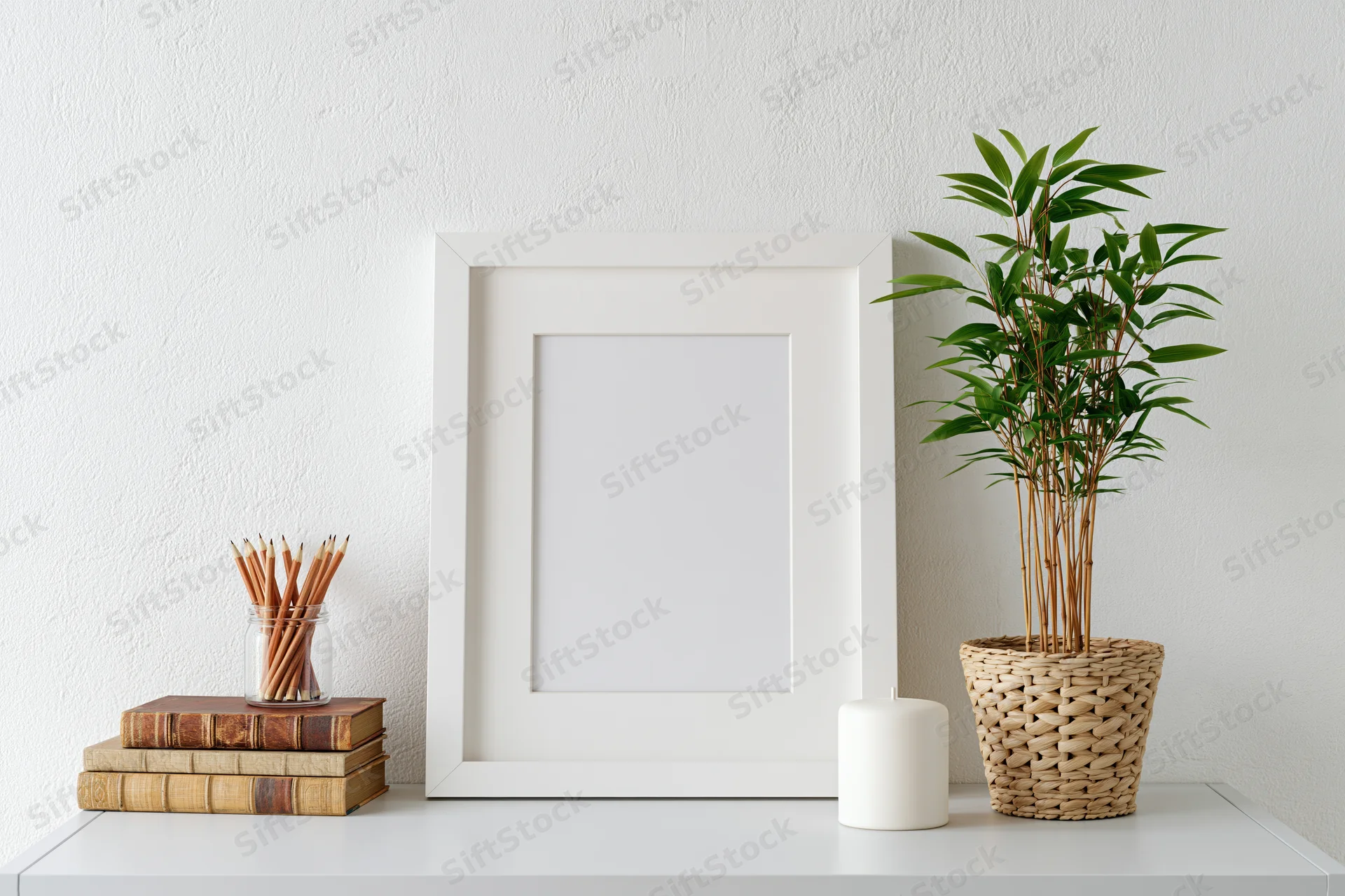 Empty white frame with wide mat leaning on white shelf beside bamboo plant in woven pot, books, pencils and candle