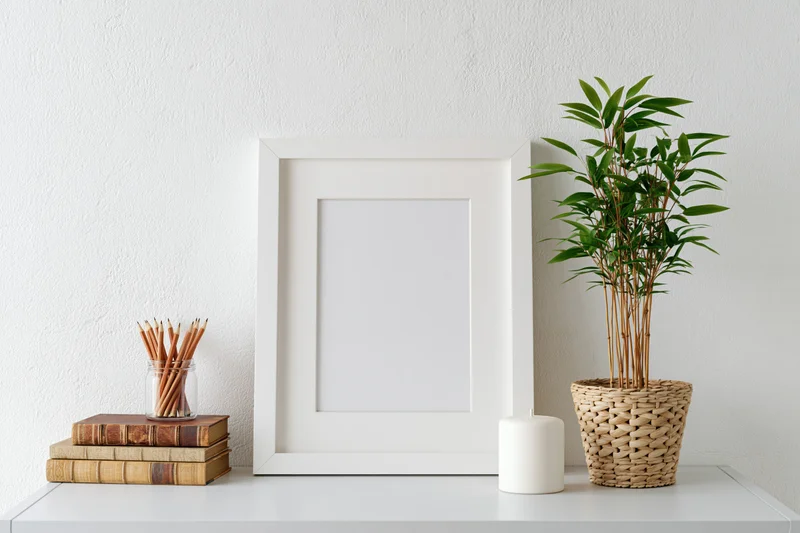 Empty white frame with wide mat leaning on white shelf beside bamboo plant in woven pot, books, pencils and candle