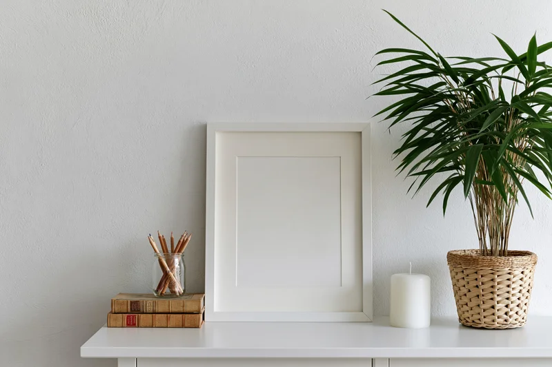 Empty white frame with wide mat leaning on white shelf beside bamboo plant in wicker pot, books, pencils and candle