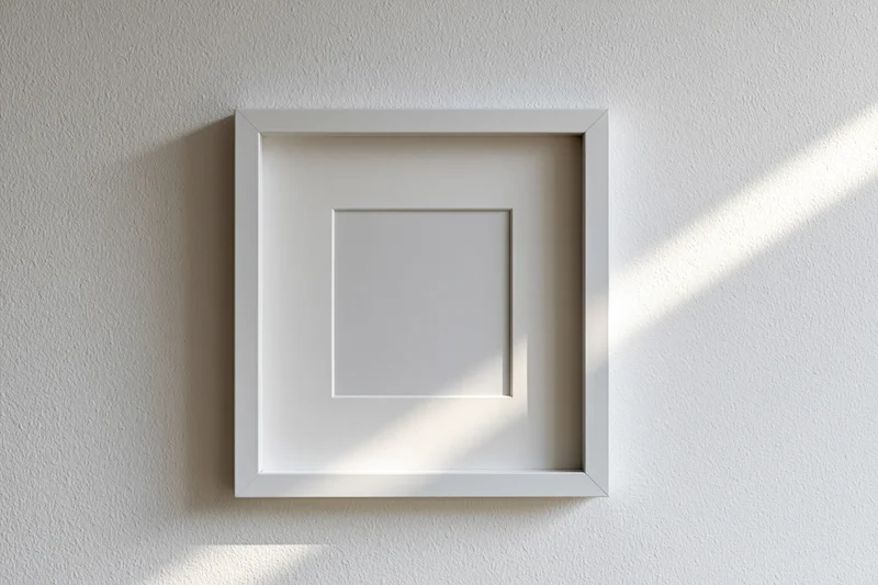 White square picture frame with wide mat hanging on textured wall, sunlight casting diagonal shadow across surface