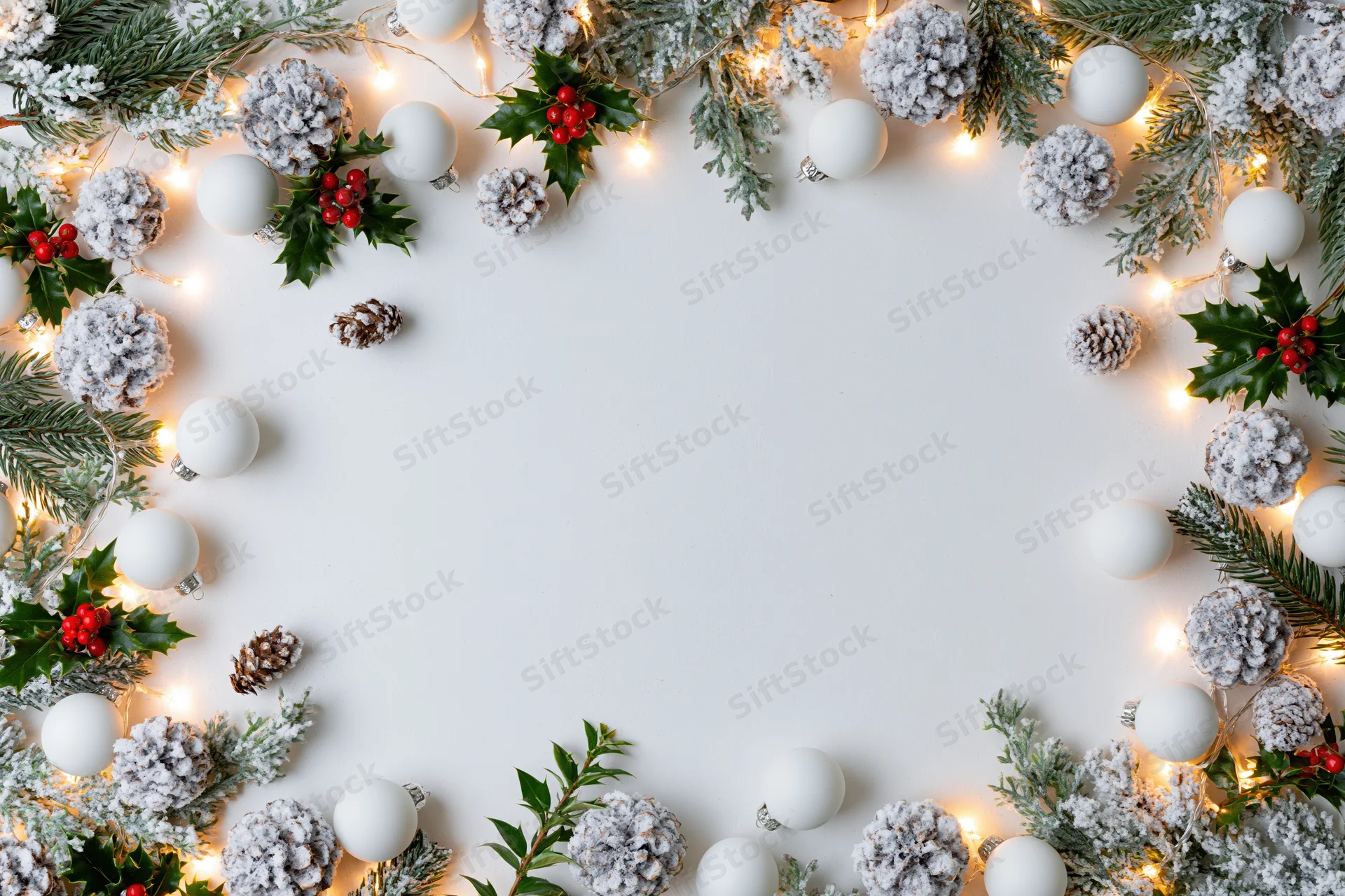 Christmas border frame of pine, holly, pinecones and lights on white surface