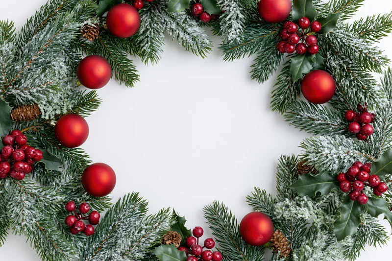 Circular christmas wreath made of pine and fir branches with red ornaments and holly berries forming a border on white background