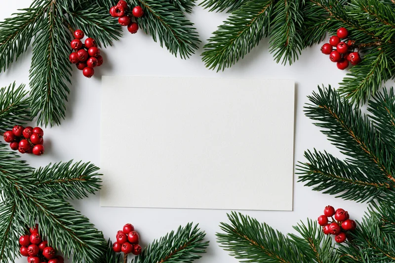 Blank white card surrounded by pine branches and red berries on white surface