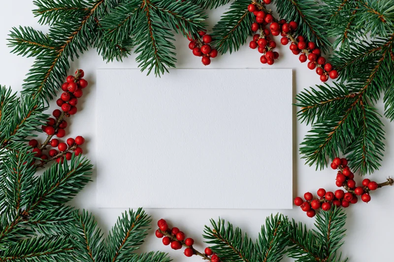 Blank white card surrounded by pine branches and red berries in a flat lay arrangement