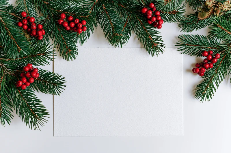 Blank white card surrounded by pine branches and red berries in a flat lay arrangement