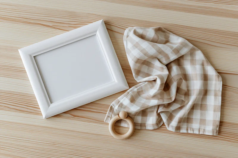 White wooden frame with blank interior and baby teether on checkered cloth on light wood surface