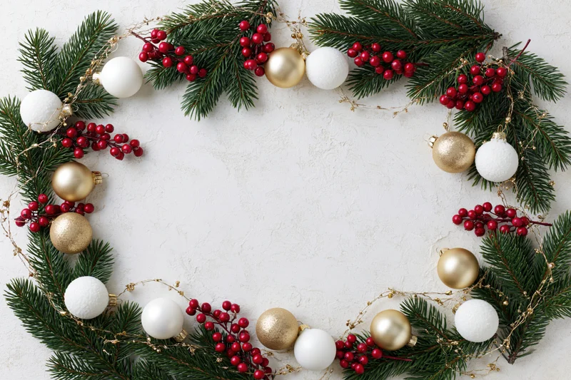 Circular wreath made of pine branches, red berries, and gold and white ornaments arranged on a textured white surface