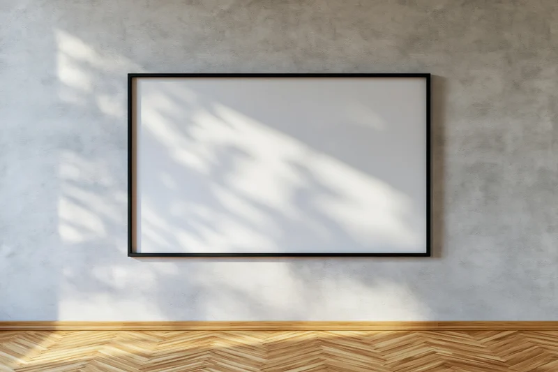 Black-framed blank canvas hanging on a concrete wall with sunlight casting shadows on a wooden parquet floor