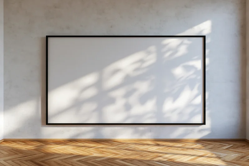 Black-framed empty canvas hanging on concrete wall in minimalist room with sunlight casting tree shadows on wooden floor