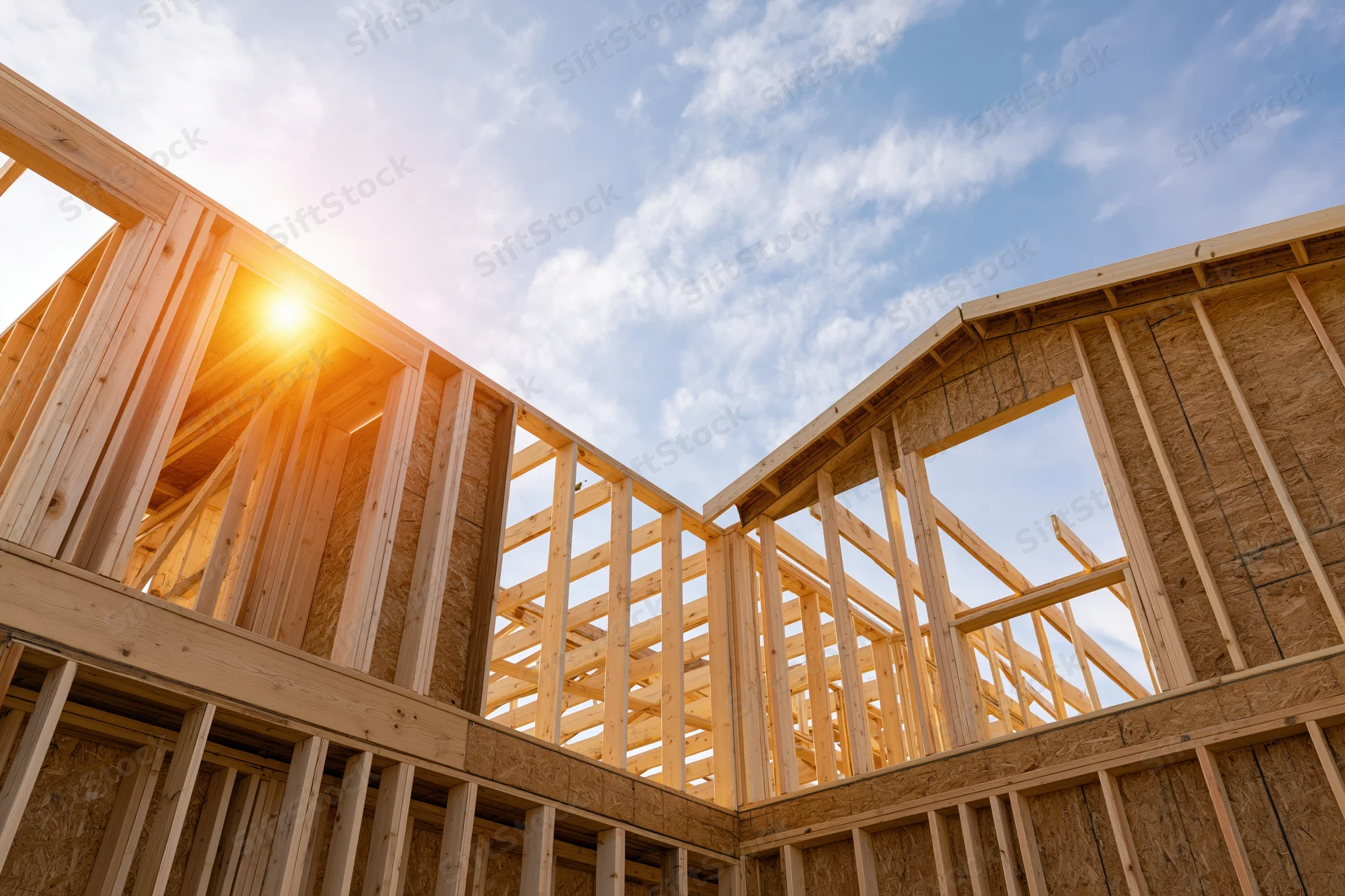 Wooden frame of a new residential house under construction against a bright blue sky with sunlight shining through the beams