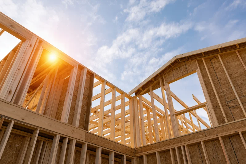 Wooden frame of a new residential house under construction against a bright blue sky with sunlight shining through the beams
