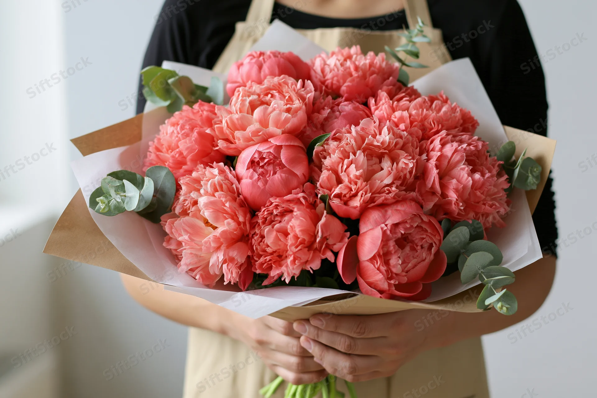 Woman holding a large bouquet of coral-pink peonies wrapped in paper with eucalyptus leaves