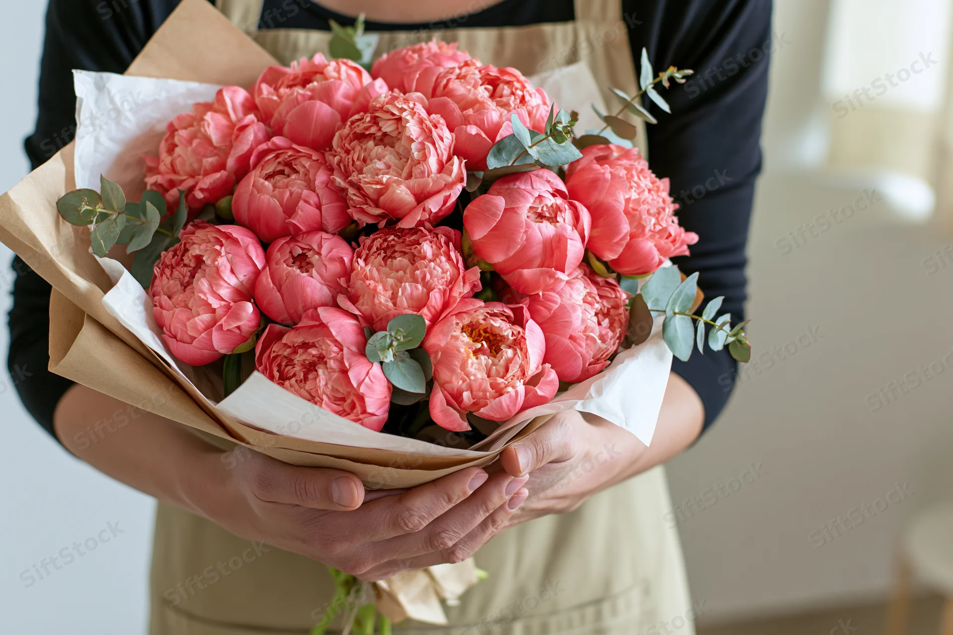 Woman holding a large bouquet of coral-pink peonies wrapped in paper with eucalyptus greenery