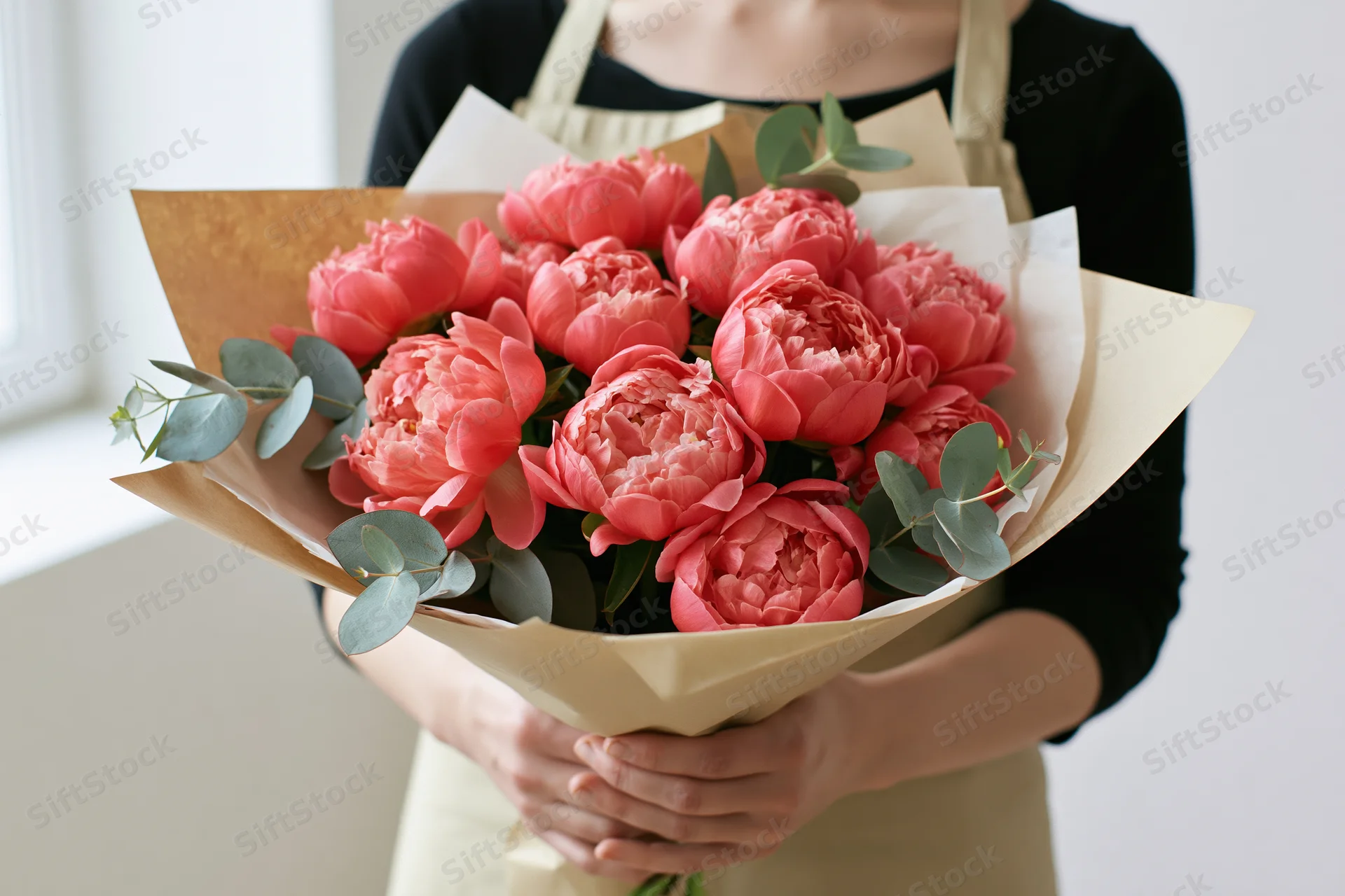 Woman holding a large bouquet of coral-pink peonies wrapped in paper with eucalyptus leaves