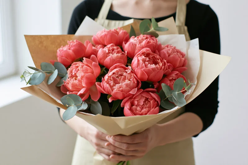 Woman holding a large bouquet of coral-pink peonies wrapped in paper with eucalyptus leaves