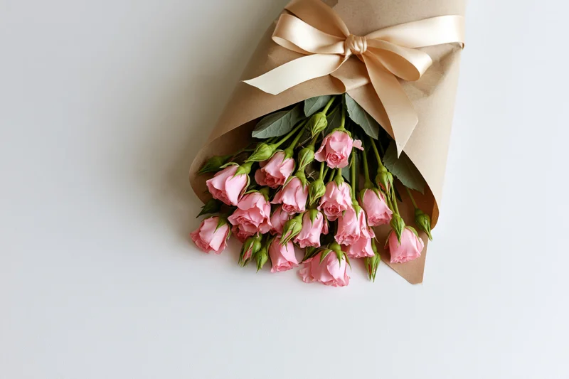 Small bouquet of pink spray roses wrapped in kraft paper with beige ribbon on white surface