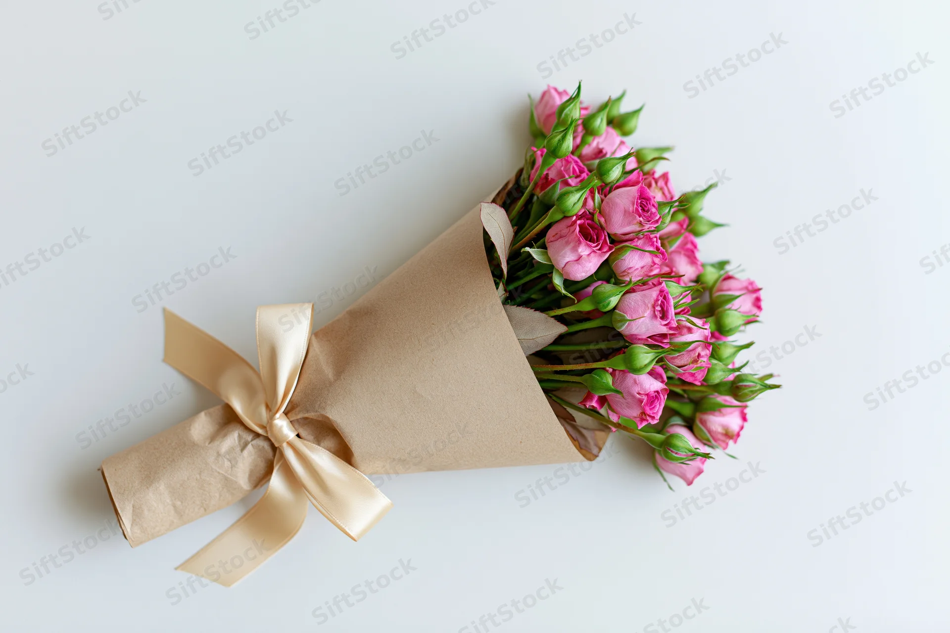Small bouquet of pink spray roses and green buds wrapped in brown paper with cream ribbon on white surface